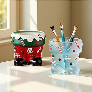 May include: Two festive pen holders in the shape of Santa's boots. One is red with white snowflakes and green trim, the other is light blue with white snowflakes and candy cane details. Brushes are inside the blue holder.