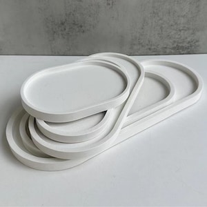 以下が含まれることがあります： 積み重ねられた4つの白い楕円形のトレイのセット。トレイは滑らかでマットな素材でできており、わずかに盛り上がった縁があります。