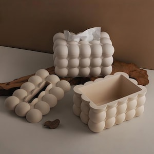 Può includere: Tre contenitori decorativi beige a forma di bolla. Uno contiene fazzoletti, un altro è una scatola rettangolare e il terzo è un coperchio. I contenitori sono disposti su una superficie chiara con un accento di legno sullo sfondo.