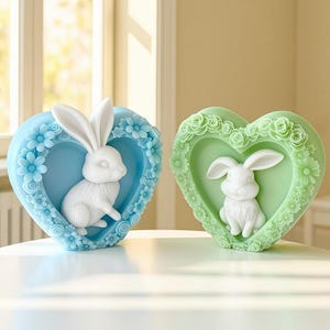 May include: Two heart-shaped decorative pieces. One is light blue with a white bunny inside, surrounded by flowers. The other is green with a white bunny inside, surrounded by flowers. Both are on a white surface.