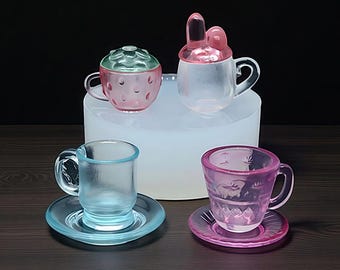 Könnte beinhalten: Vier farbige Teetassen und Untertassen aus Glas. Zwei Teetassen haben Deckel, einer in Erdbeerform und der andere mit Hasenohren. Die anderen beiden Teetassen sind blau und rosa, mit passenden Untertassen. Die Tassen stehen auf einer dunklen Holzoberfläche.