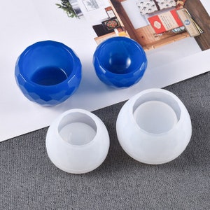 Peut inclure: Quatre moules en silicone pour la fabrication d'objets en résine. Deux moules sont bleus et deux sont blancs. Les moules sont ronds et ont une lèvre légèrement surélevée autour du haut.