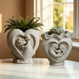 May include: Two heart-shaped planters with detailed relief carvings. One features a woman holding flowers, the other a mother and child. The planters are filled with greenery and a succulent, and are made of a light-colored stone.