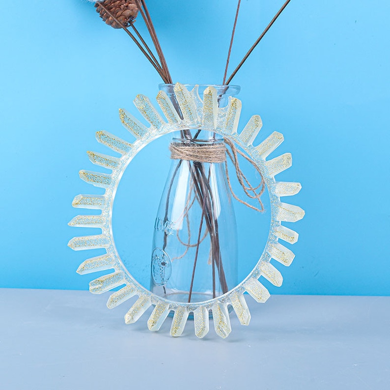 May include: A clear resin ring with a gold glitter finish. The ring is shaped like a sun with many points. The ring is sitting on a blue surface.