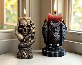 May include: Two lit decorative candles. One is shaped like a skull held by a hand with a snake coiled around the base. The other is a dark red owl candle on a black geometric base. Both candles are on a white surface.