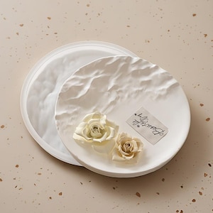 May include: Two white, textured, round decorative trays. One tray is partially visible, while the other is in the foreground, adorned with two cream-coloured roses and a tag that reads "Beautiful Day".