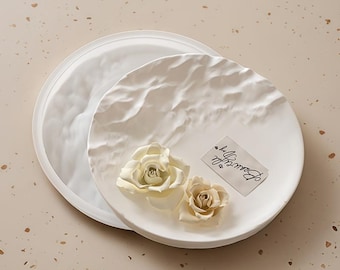 Può includere: Due vassoi decorativi rotondi bianchi e strutturati. Un vassoio è parzialmente visibile, mentre l'altro è in primo piano, adornato con due rose color crema e un'etichetta con la scritta "Beautiful Day".