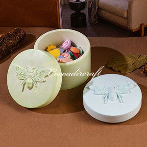 Puede incluir: Dos cajas redondas con tapa y diseños de abejas. La tapa de una caja es verde claro con una abeja blanca, y la tapa de la otra caja es azul claro con una abeja blanca. La caja con la tapa verde está abierta, revelando una pequeña colección de botones y cuentas de colores.