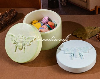 May include: Two round, lidded boxes with bee designs. The lid of one box is light green with a white bee, and the lid of the other box is light blue with a white bee. The box with the green lid is open, revealing a small collection of colourful buttons and beads.