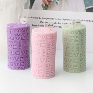 May include: Three cylindrical candles with the word "LOVE" embossed on them. The candles are purple, pink, and green.