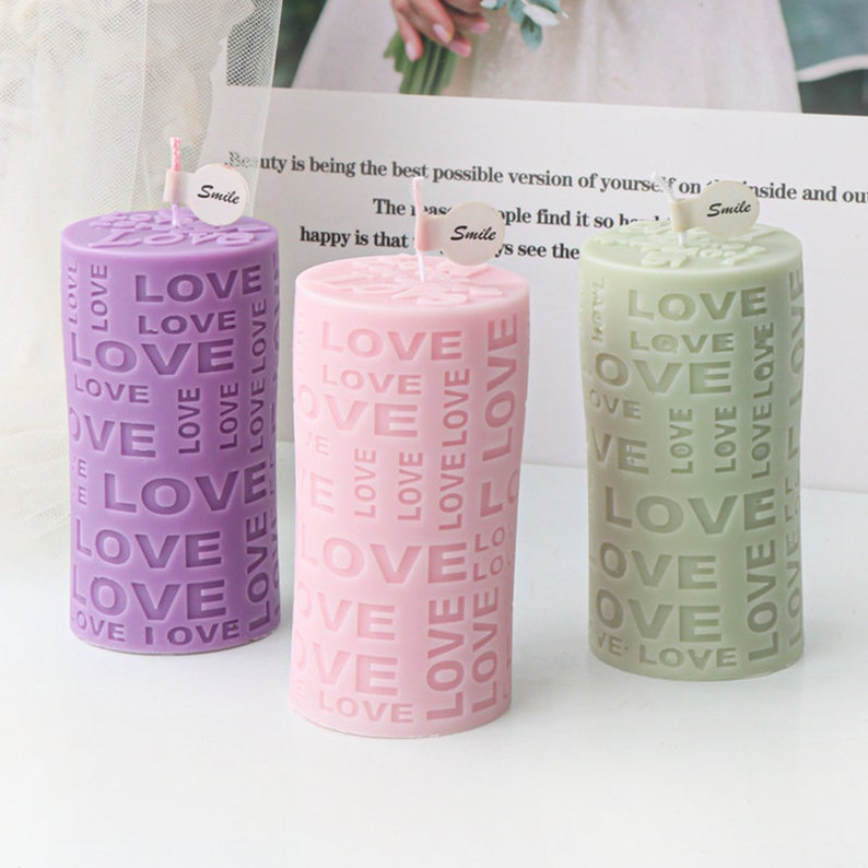 May include: Three cylindrical candles with the word "LOVE" embossed on them. The candles are purple, pink, and green.