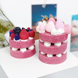 May include: Two miniature pink cakes decorated with frosting and various colorful candies. The cake on the left is topped with blackberries and strawberries. The cake on the right is topped with pink and white candies, chocolate, and a swirl of pink frosting.