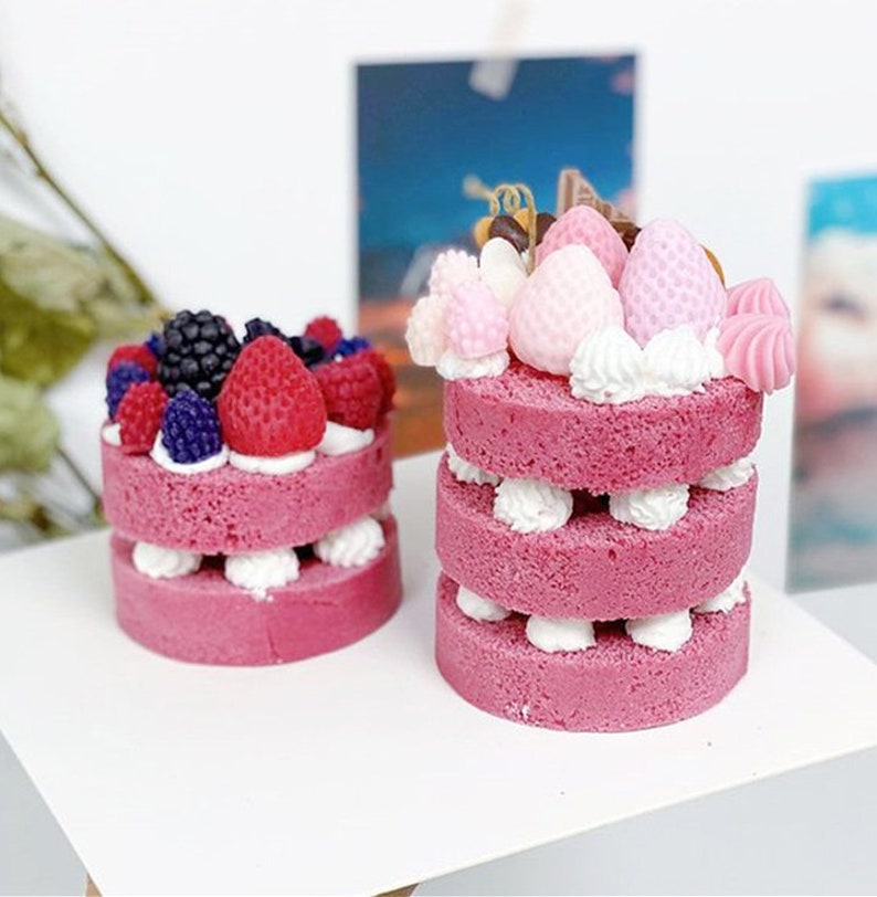 May include: Two miniature pink cakes decorated with frosting and various colorful candies. The cake on the left is topped with blackberries and strawberries. The cake on the right is topped with pink and white candies, chocolate, and a swirl of pink frosting.