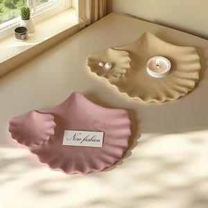 Könnte beinhalten: Zwei dekorative Muschelschalen. Eine ist hellrosa, mit einer kleinen weißen Karte mit der Aufschrift "Now fashion". Die andere ist beige, mit einer brennenden Kerze und zwei Perlen. Die Schalen stehen auf einer hellbeigen Oberfläche.