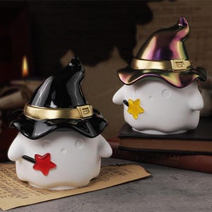 May include: Two white ghost figurines with black and iridescent witch hats. Each ghost holds a star-shaped wand. The ghost on the left has a red wand and the ghost on the right has a yellow wand.