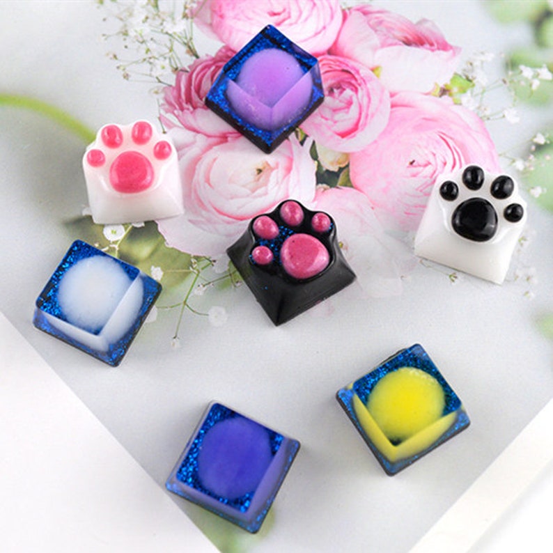 May include: Set of 7 colorful keycaps with paw print designs. The keycaps are made of resin and have a glossy finish. The colors include purple, pink, black, white, and yellow.