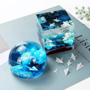 Peut inclure: Un cube et une sphère en résine transparente remplis de résine bleue et de petits avions en papier blanc. Le cube et la sphère sont sur une surface bleu clair avec des petits avions en papier blanc éparpillés autour d'eux.