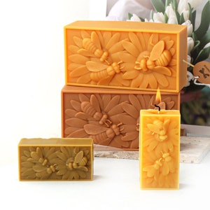 May include: Three beeswax candles with a bee and flower design. The candles are in different sizes and are arranged in a pyramid shape. The largest candle is yellow and the two smaller candles are brown.
