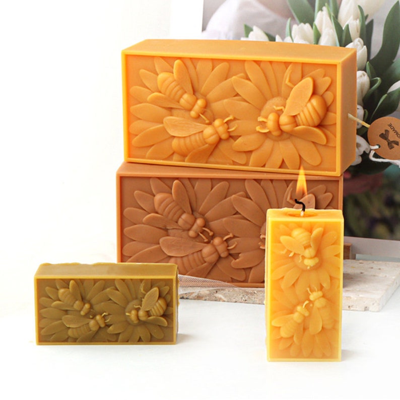 May include: Three beeswax candles with a bee and flower design. The candles are in different sizes and are arranged in a pyramid shape. The largest candle is yellow and the two smaller candles are brown.