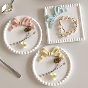 Puede incluir: Platos decorativos de cerámica blanca con bordes realzados y lazos en rosa, amarillo y azul. Cada plato presenta un diseño floral con una flor seca y una pequeña flor blanca. Un plato cuadrado contiene un collar de perlas.