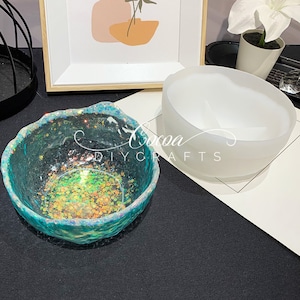 May include: Two resin bowls, one clear with iridescent glitter and a teal rim, and one white, on a black surface. The text "Cocoa DIY Crafts" is visible in the image.