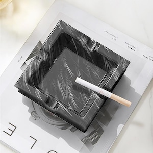 Peut inclure: Un cendrier carré noir avec un motif blanc en forme de plumes. Une cigarette allumée repose dans le cendrier. Le cendrier est posé sur un magazine avec du texte.