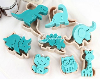 May include: A collection of teal-colored animal-shaped candles, including a Tyrannosaurus Rex, a Brontosaurus, a Triceratops, a Stegosaurus, an elephant, a frog, an owl, and a giraffe. Each candle has a wick and is set in a light brown mold.