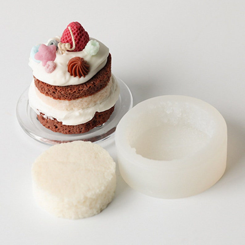 May include: A white silicone cake mold with a round, shallow base. The mold is designed to create a small, layered cake. A finished cake is shown next to the mold, decorated with frosting and colorful candies.
