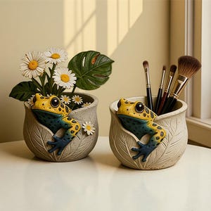 Può includere: Due vasi in ceramica a forma di rana. Un vaso contiene margherite e una grande foglia verde. L'altro vaso contiene pennelli per il trucco. Le rane sono gialle e blu con macchie nere e i vasi sono grigio chiaro.