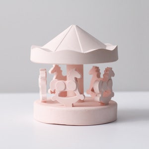 May include: A pink miniature carousel with four horses, a roof, and a base. The carousel is made of a smooth, matte material.