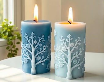 May include: Two lit blue pillar candles with white tree and snowflake designs. The candles have a cylindrical shape and are placed on a white surface. The flames are bright yellow.