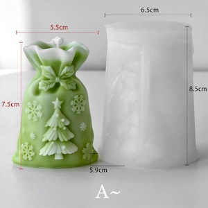 May include: A green Christmas-themed candle shaped like a gift bag, 7.5 cm tall and 5.5 cm wide, with a white bow, snowflakes, and a Christmas tree design. Next to it is a white, cylindrical mold, 8.5 cm tall and 6.5 cm wide.