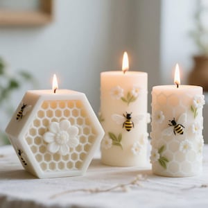 May include: Three lit, ivory-colored candles. One is hexagonal with a honeycomb design and a flower, the others are cylindrical with bees, flowers, and honeycomb patterns. The candles are on a white surface.