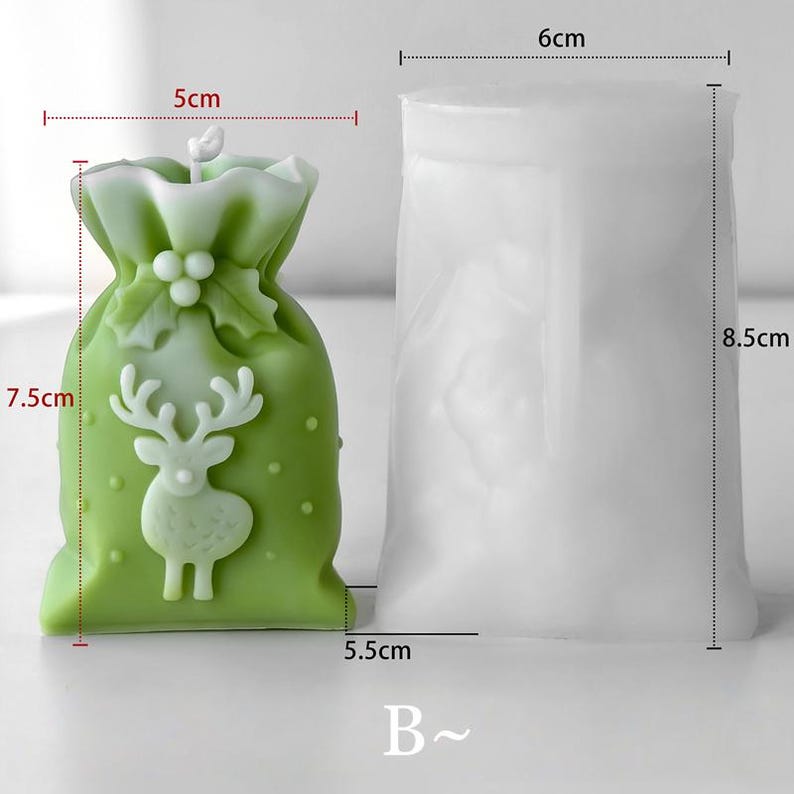 May include: A green candle in the shape of a gift bag, 7.5 cm tall and 5 cm wide, with a white reindeer design. Beside it is a white silicone mould, 8.5 cm tall and 6 cm wide, for candle making. The candle has a festive theme.
