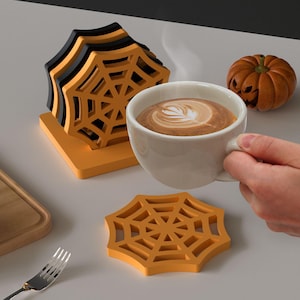 May include: A stack of spiderweb-shaped coasters in black and orange, with a single coaster in front. A white coffee cup with latte art is held by a hand, and a carved pumpkin sits nearby. Halloween-themed home decor.