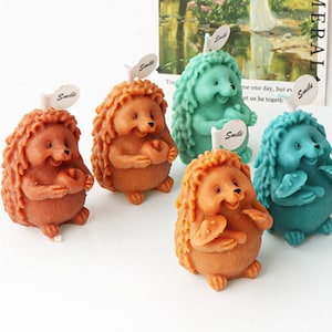 May include: Four hedgehog-shaped candles, two brown, one teal, and one light green, with a smile on their faces and holding a small heart in their paws. Each candle has a small tag that says "Smile".