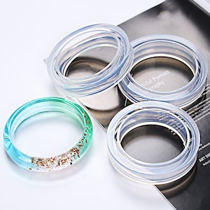Peut inclure: Trois moules en silicone transparents pour la fabrication de bracelets en résine. Les moules sont empilés les uns sur les autres. Un bracelet en résine fini avec des accents verts et dorés est également présenté.