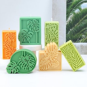 Puede incluir: Una colección de velas decorativas en varias formas y colores, incluyendo naranja, verde y amarillo. Cada vela presenta un diseño detallado de hojas y una mecha blanca en la parte superior. Las velas están dispuestas sobre una superficie blanca.