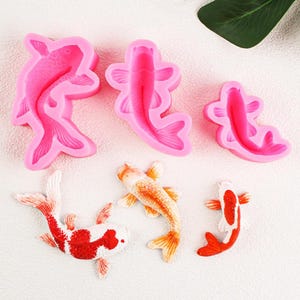 Könnte beinhalten: Ein Set mit drei rosa Silikonformen in Form von Koi-Fischen und zwei kleinen Koi-Fischen aus Fondant oder Gummipaste. Die Formen werden verwendet, um essbare Dekorationen für Kuchen und andere Desserts zu erstellen.