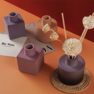 May include: An assortment of decorative ceramic essential oil diffusers in shades of purple and peach. One diffuser has a woven coaster and decorative reeds and flowers. The text "Be You" is visible on a card.