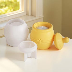 Puede incluir: Una colección de portavelas decorativos. Incluye un portavelas redondo blanco texturizado, un pequeño portavelas blanco y un portavelas amarillo con forma de tarro de miel con un diseño de abeja. El tarro de miel tiene una tapa con la palabra "HONEY".