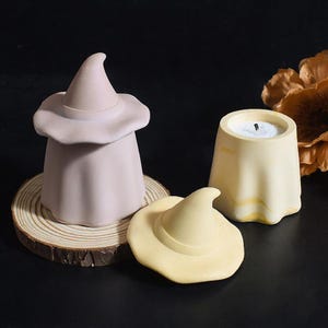 Könnte beinhalten: Eine Sammlung von Halloween-Kerzen und Dekorationen. Das Bild zeigt einen hellvioletten Hexenhut-Kerzenhalter, einen gelben Hexenhut und einen gelben Kerzenhalter mit einer brennenden Kerze. Die Artikel sind auf einer dunklen Oberfläche angeordnet.