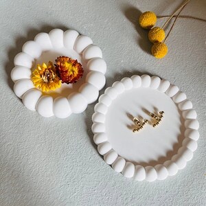 Peut inclure: Deux plateaux ronds blancs avec un bord en forme de bulles. L'un des plateaux contient deux fleurs jaunes séchées, et l'autre une paire de boucles d'oreilles dorées. Quelques boules décoratives jaunes et des fleurs séchées sont en arrière-plan.