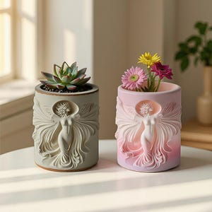May include: Two decorative ceramic planters, one gray and one pink, each featuring a sculpted angel design. The gray planter holds a succulent, while the pink one contains colorful flowers. The planters are cylindrical and sit on a white surface.
