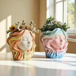 Può includere: Due vasi decorativi a forma di volti femminili sereni. Un vaso presenta un gradiente di giallo, arancione e pesca, mentre l'altro mostra tonalità rosa e blu. Entrambi i vasi sono riempiti di verde e fiori, aggiungendo un tocco naturale.