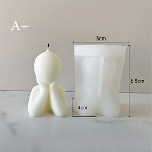 May include: A cream-colored candle in the shape of a person with their hands on their face. The candle is next to a clear silicone mold. The mold is 5 cm wide, 6.5 cm tall, and 4 cm deep. The letter "A" is in the upper left corner.