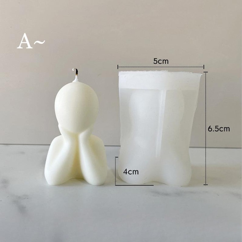 May include: A cream-colored candle in the shape of a person with their hands on their face. The candle is next to a clear silicone mold. The mold is 5 cm wide, 6.5 cm tall, and 4 cm deep. The letter "A" is in the upper left corner.