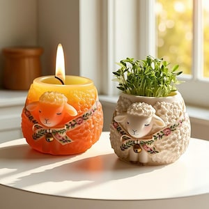 以下が含まれることがあります： 羊の形をした、火の灯ったオレンジ色のろうそくと白いプランター。 ろうそくには羊の顔と花の飾りリボンが付いています。 プランターには緑色の新芽が植えられており、羊の顔と花の飾りリボンも付いています。 どちらにも小さなベルが付いています。