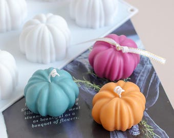 Peut inclure: Quatre bougies en forme de citrouille de différentes couleurs : blanc, bleu sarcelle, rose et orange. Les bougies sont posées sur une surface blanche avec un livre en dessous. Les bougies sont toutes de la même taille et de la même forme, et elles ont une texture côtelée.