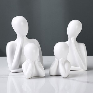 May include: Four white ceramic sculptures depicting abstract human figures. Two larger figures stand behind two smaller figures, all with a minimalist design. The sculptures have smooth surfaces and are set against a neutral background.